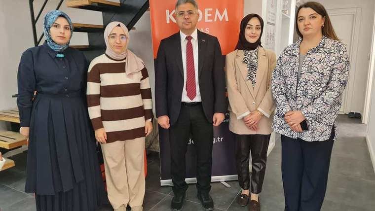 İç mekânda, üzerinde KADEM yazılı turuncu pano önünde dört kadın ve bir erkek yan yana duruyor.