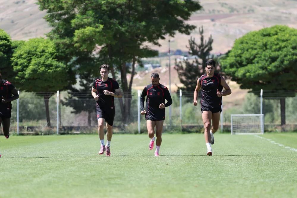 Çim sahada siyah spor kıyafetleri giyen dört kişi koşu çalışması yapıyor; arkada kale ve ağaçlar görülüyor.