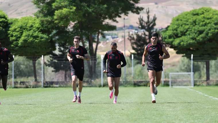 Çim sahada siyah spor kıyafetleri giyen dört kişi koşu çalışması yapıyor; arkada kale ve ağaçlar görülüyor.