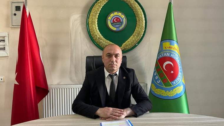 Masanın arkasında oturan bir kişi, Türk bayrağı ve amblemli yeşil bayrağın arasında duruyor.
