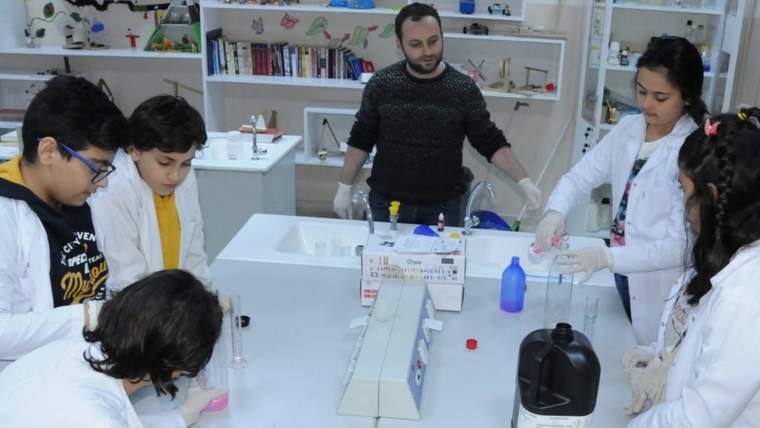 Laboratuvar ortamında deney yapan öğretmen ve öğrenciler, masanın etrafında toplanmış.