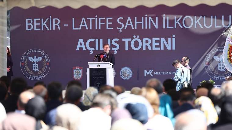 Üzerinde “Bekir-Latife Şahin İlkokulu Açılış Töreni” yazan sahnede kürsüde konuşan kişi ve kalabalık