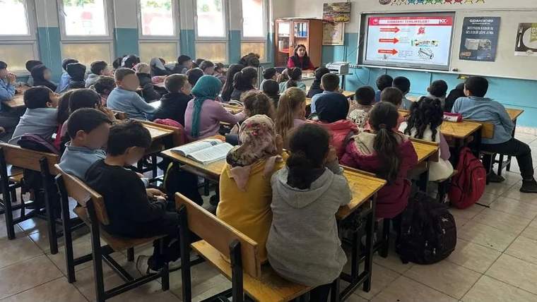 Sınıfta sıralarda oturan öğrenciler, önde sunum yapan öğretmeni ve ekrandaki görselleri izliyor.