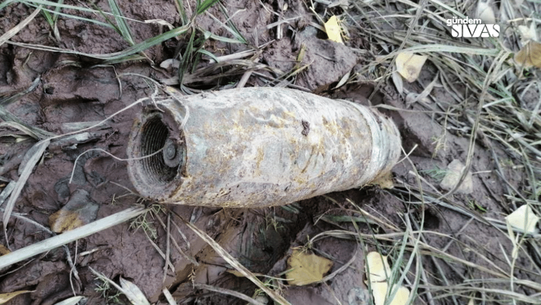 Toprak ve kuru otlar arasında, paslanmış silindirik metal bir nesne yan yatmış halde duruyor.