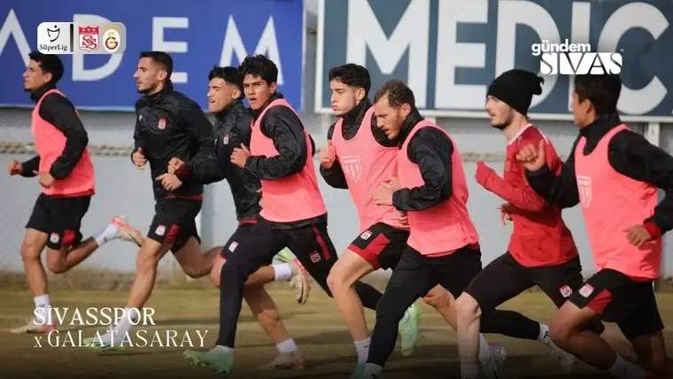 Çim sahada pembe yelekli futbolcular, yanlarında siyah eşofmanlı oyuncularla birlikte koşuyor.