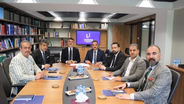 Bir toplantı odasında, etrafında yedi kişi bulunan bir masa yer alıyor. Masada not defterleri ve atıştırmalıklar var.