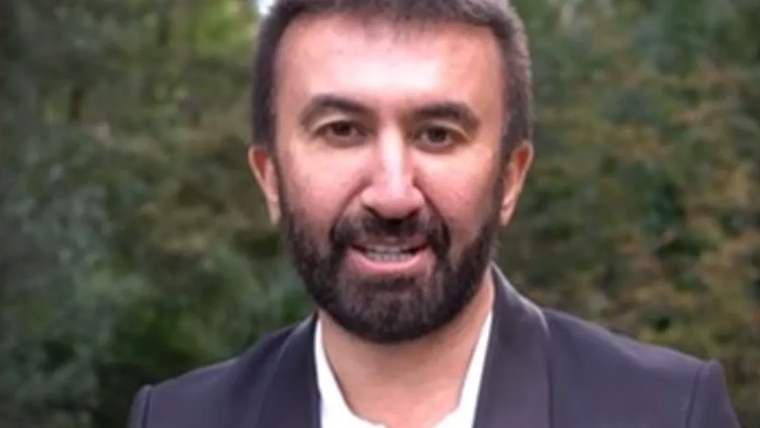 Kahverengi sakallı bir adam, siyah ceket giymiş, gülümseyerek kameraya bakıyor.