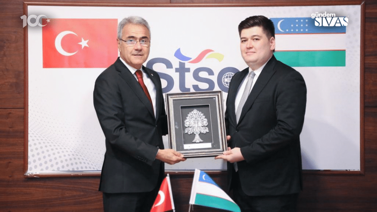 Takım elbiseli iki kişi, çift başlı kartal motifli plaketi tutuyor; arkada Türk ve Özbekistan bayrakları var.