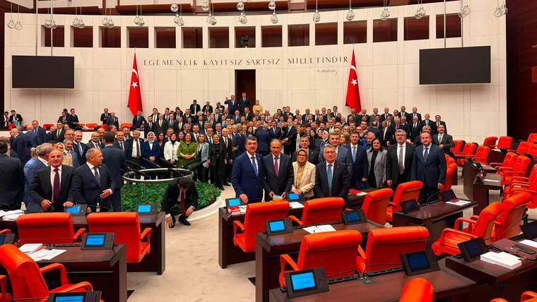 Kırmızı koltuklu geniş salonda, Türk bayrakları önünde çok sayıda kişi toplu halde duruyor.