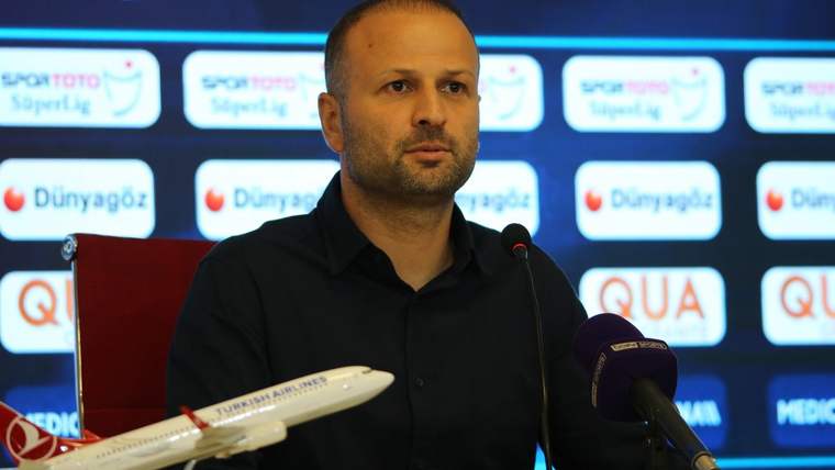 Basın toplantısında konuşan bir kişi, önünde mikrofonlar ve sponsor logoları var.