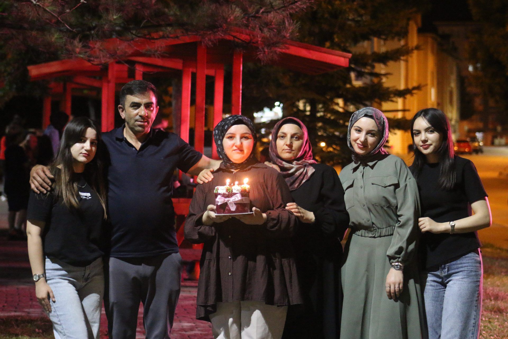 Gece dışarıda altı kişi, mumları yanan pastayı tutan genç kadının çevresinde yan yana duruyor.