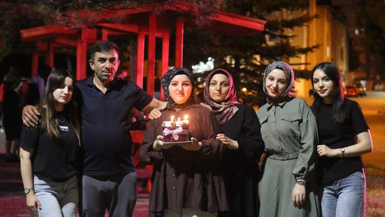 Gece dışarıda altı kişi, mumları yanan pastayı tutan genç kadının çevresinde yan yana duruyor.