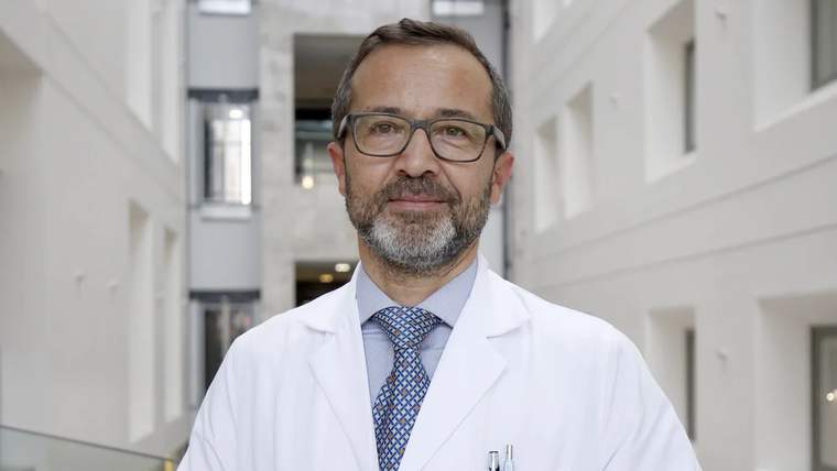 Beyaz önlük giymiş bir doktor, modern bir binanın içinde gülümseyerek poz veriyor.