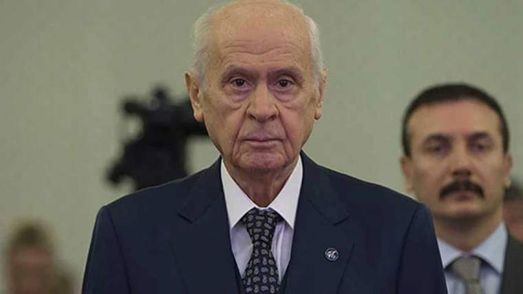 Koyu takım elbiseli yaşlı bir erkek önde duruyor; arkasında takım elbiseli başka bir erkek ve kamera görülüyor.