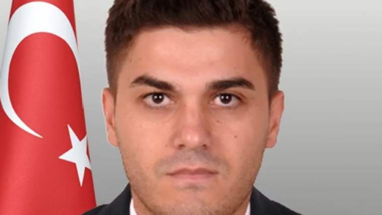 Koyu renk takım elbiseli bir erkek, arka planda Türk bayrağı bulunan bir portrede kameraya bakıyor.