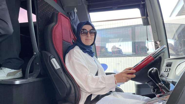 Otobüs koltuğunda oturan bir kadın, direksiyonu tutuyor ve güneş gözlüğü takıyor.
