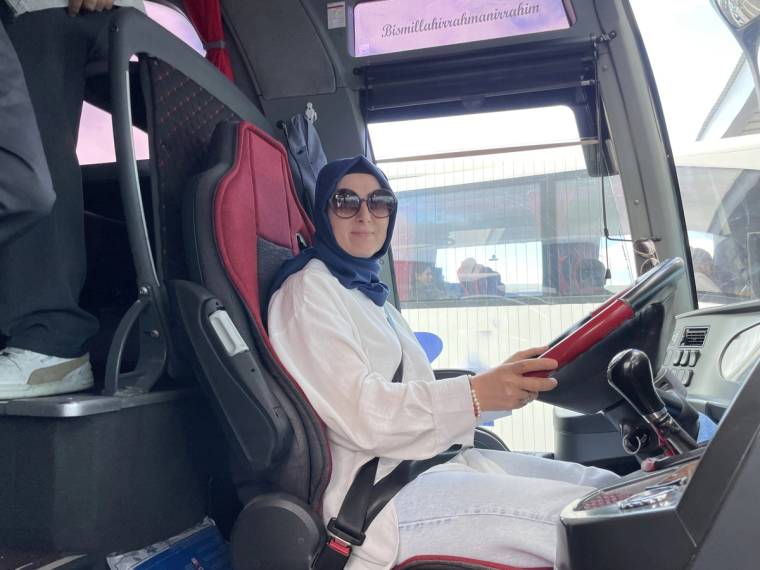 Otobüs koltuğunda oturan bir kadın, direksiyonu tutuyor ve güneş gözlüğü takıyor.