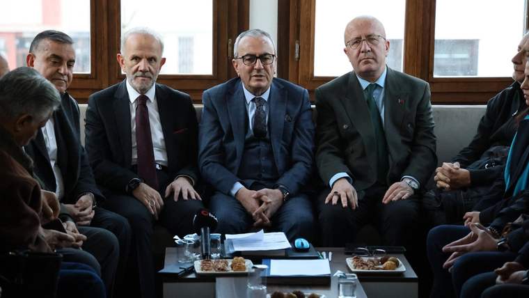 Bir grup erkek, bir odada oturuyor; masada belgeler ve atıştırmalıklar var.