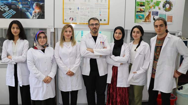 Laboratuvar ortamında beyaz önlük giyen yedi kişi, duvarlardaki bilimsel afişlerin önünde yan yana duruyor.