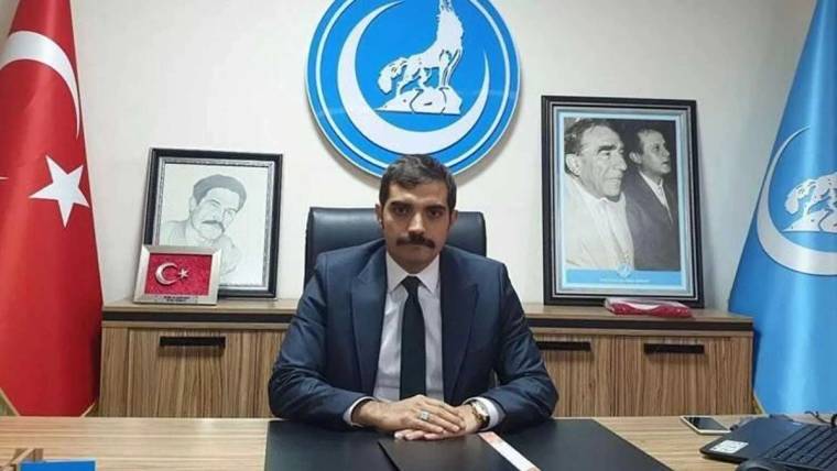 Bıyıklı bir kişi, Türk bayrağı ve mavi bayrakların yanında, amblem ile çerçeveli portrelerin önünde oturuyor.