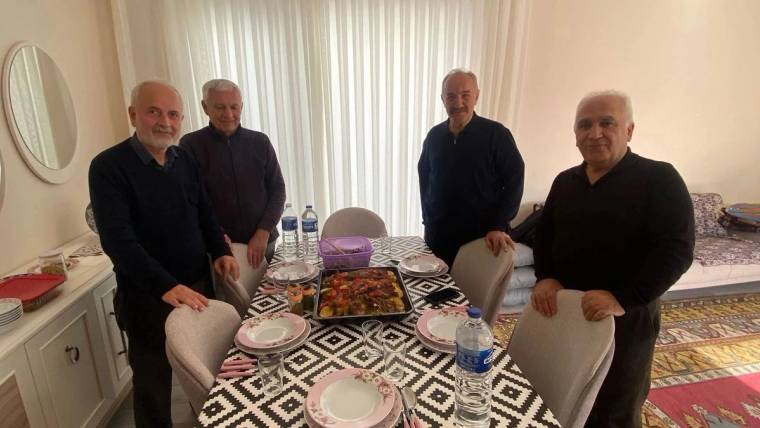 Aydınlık bir odada dört erkek, yemek ve su şişelerinin bulunduğu masanın çevresinde birlikte duruyor.