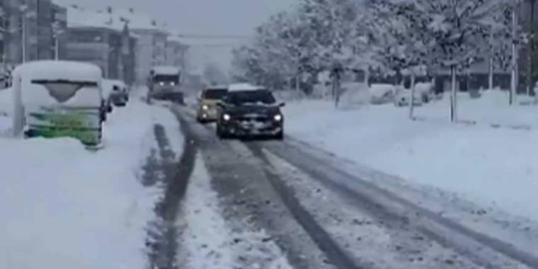 Kar yağışı altında ilerleyen otomobiller ve park etmiş araçların bulunduğu karlı cadde, ağaçlarla çevrili.