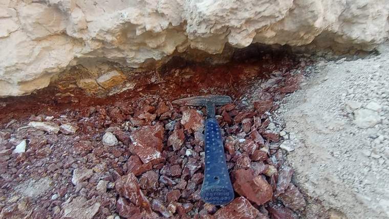 Kırmızı taşların arasında bir kazı aleti yer alıyor, arka planda kayalık bir yüzey görünüyor.