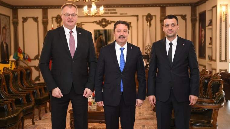 Üç kişi, klasik bir ofis ortamında yan yana duruyor; arka planda Türk bayrağı ve diğer bayraklar görünmekte.