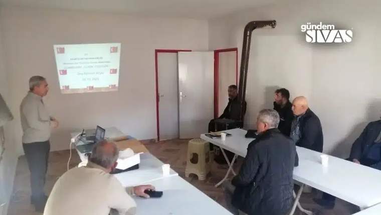 Bir odada, duvardaki sunum perdesinin önünde konuşan kişi ve masalarda oturan yetişkinler görülüyor.