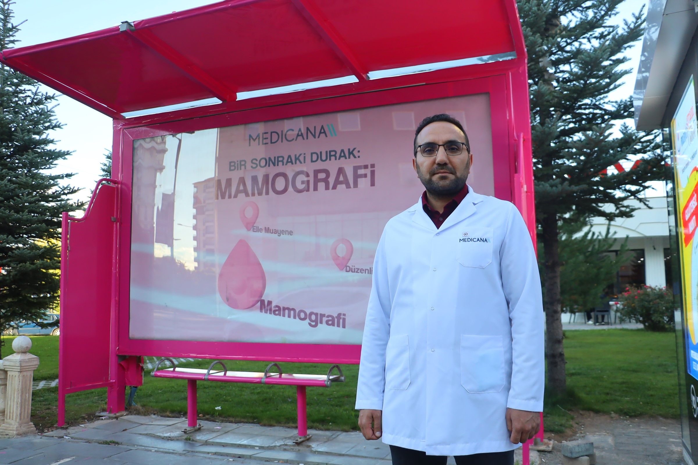 Gözlüklü bir kişi beyaz önlükle, üzerinde “Bir Sonraki Durak: Mamografi” yazan pembe durak yanında duruyor.