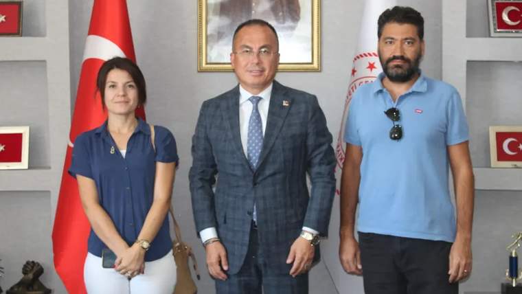 Türk bayrağı önünde, biri takım elbiseli üç kişi bir odada yan yana duruyor.