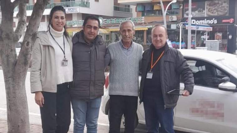 Kaldırımda dört kişi, beyaz bir otomobilin önünde yan yana duruyor; arkada mağaza tabelaları görülüyor.