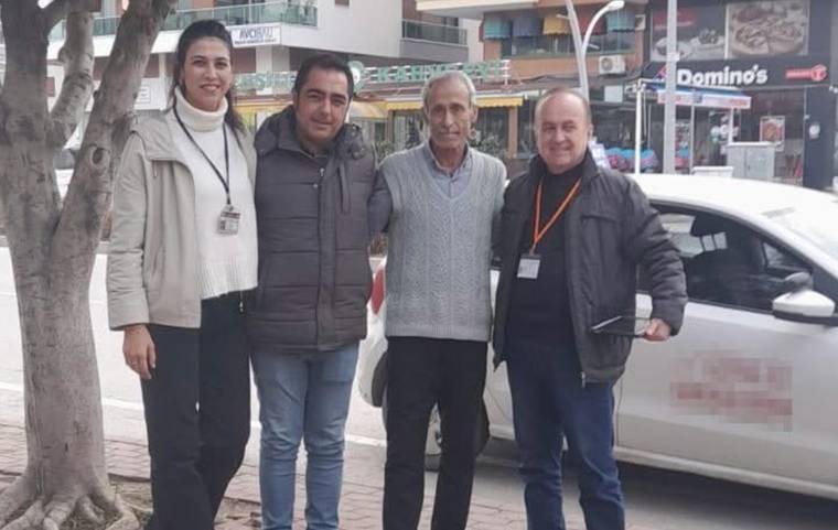 Kaldırımda dört kişi, beyaz bir otomobilin önünde yan yana duruyor; arkada mağaza tabelaları görülüyor.