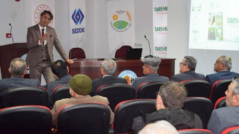 Toplantı salonunda kürsü önünde konuşan bir kişi ve onu dinleyen katılımcılar, arkada TARSİM afişleri.