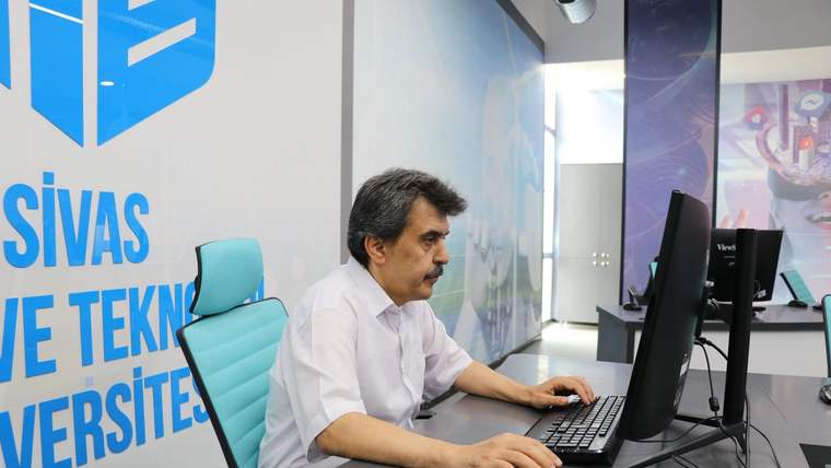 Bir kişi, duvarda üniversite yazısı bulunan ofiste iki monitörlü bilgisayar başında çalışıyor.