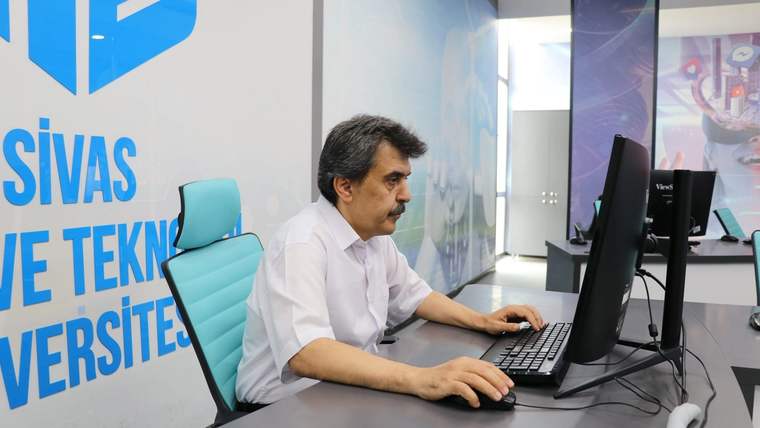 Bir kişi, duvarda üniversite yazısı bulunan ofiste iki monitörlü bilgisayar başında çalışıyor.