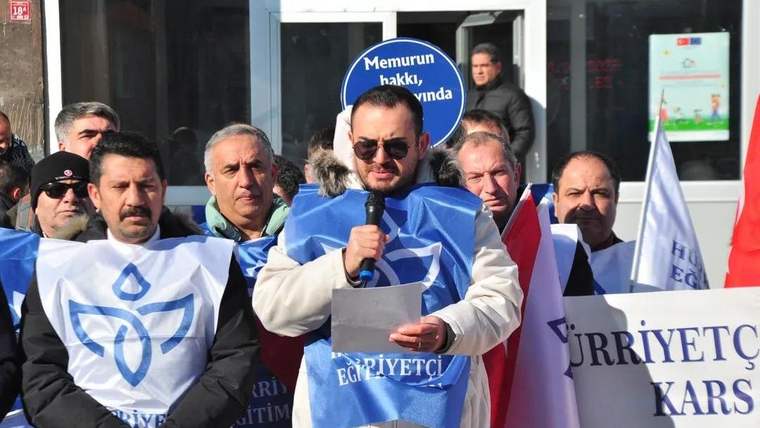 Mikrofonla konuşan bir kişi, çevresindeki kalabalık ve Hürriyetçi Eğitim Sen yazılı pankartlarla bina önünde duruyor.