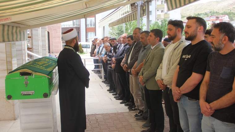 Yeşil örtüyle kaplı bir tabutun yanında cübbeli bir kişi dururken, karşısında sıra halinde erkekler bekliyor.