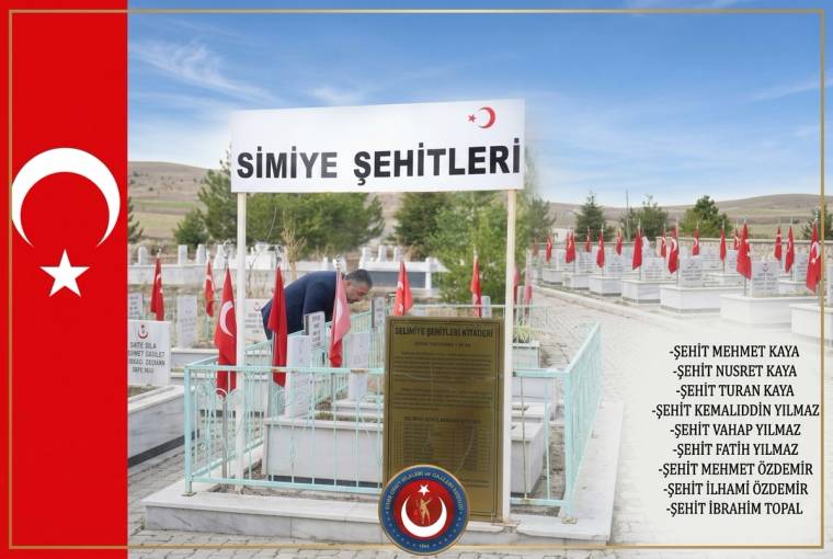 Türk bayraklarıyla çevrili şehitlikte, üzerinde “ŞEHİTLERİ” yazan tabela ve anıt plaketi görülüyor.