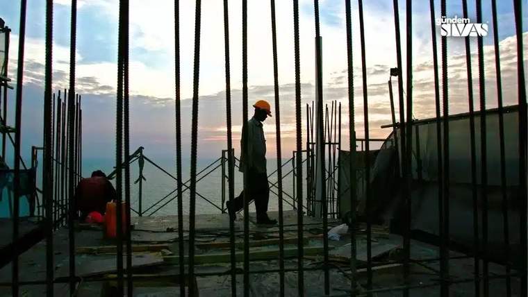 Bir inşaat alanında, işçi kask takarak yürürken, arka planda deniz ve gökyüzü görünmekte.