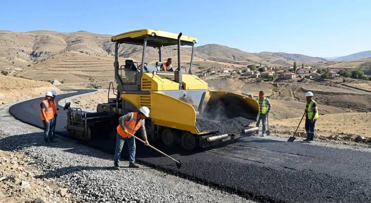 Dağlık arazide sarı asfalt finişerinin çevresinde çalışan işçiler, yeni serilen asfaltı düzenliyor.