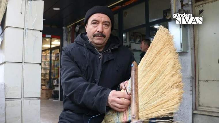 Siyah bereli bir adam, dükkân girişinde elinde saplı geleneksel bir süpürge tutuyor.