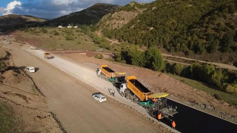 Dağlık ve ormanlık alanda, asfalt serimi yapılan yolda damperli kamyonlar ve işçiler görülüyor.