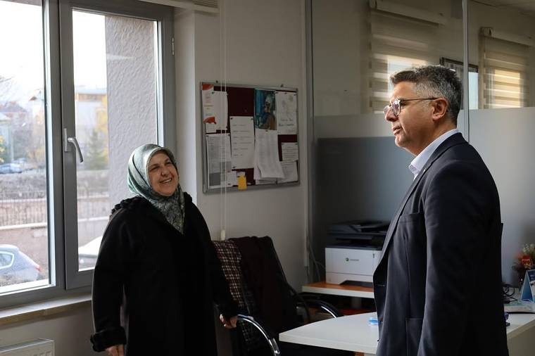 Ofis ortamında başörtülü bir kadın ile gözlüklü bir erkek, masa yanında ayakta konuşuyor.
