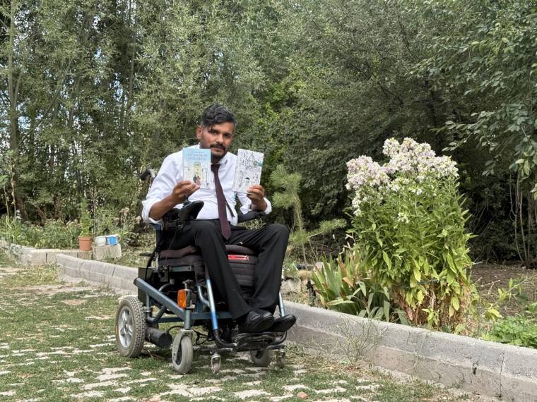 Tekerlekli sandalyede oturan bir adam, elinde iki kitapçığı tutarak ağaçlık alanda kameraya bakıyor.