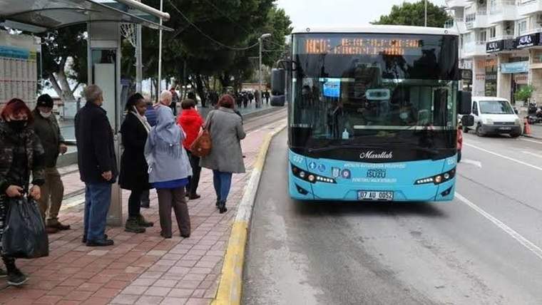 Turkuaz renkli belediye otobüsü durakta ilerlerken kaldırımda bekleyen yolcular görülüyor.