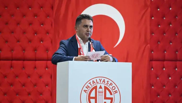 Kürsüde konuşan bir kişi, arkasındaki Türk bayrağı önünde Antalyaspor logolu kürsüde duruyor.