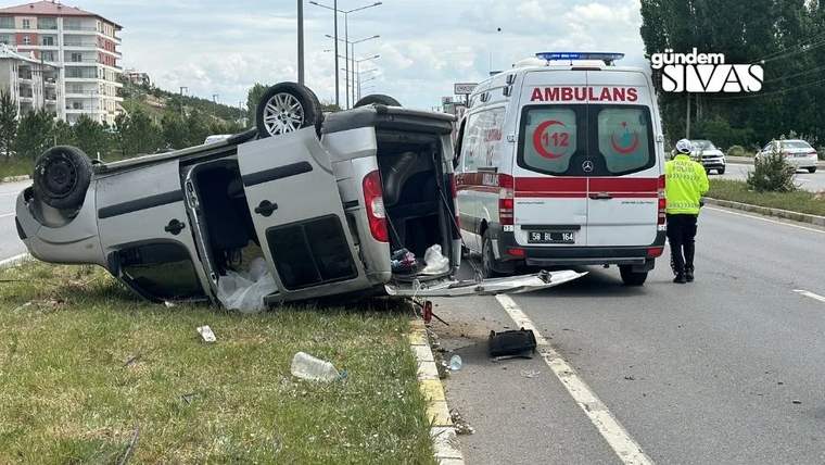 Ters dönen bir hafif ticari araç yol kenarında, arkasında ambulans ve yanında reflektif yelekli kişi bulunuyor.