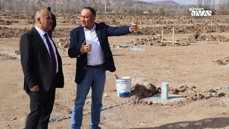 Kurak bir arazide duran iki erkekten biri elini uzatırken diğeri yanında bekliyor; yerde hortum ve kova görülüyor.