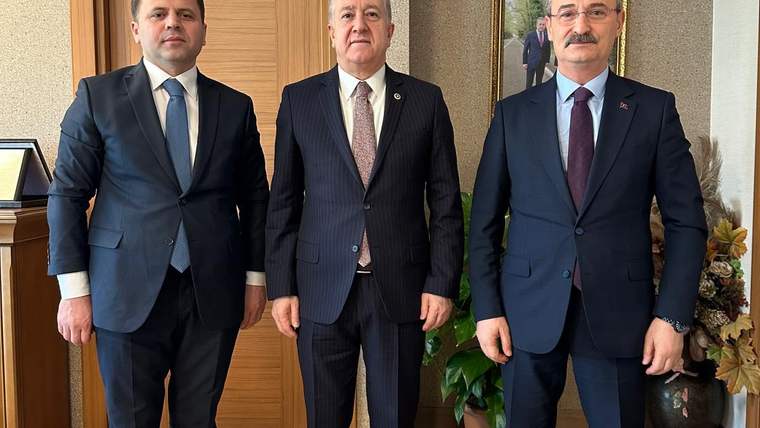 Takım elbiseli üç kişi, ahşap kapı önünde yan yana ayakta duruyor; arkalarında tablo ve bitki bulunuyor.
